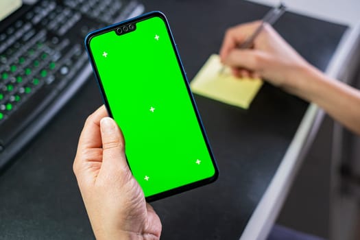 Woman hands hold a smartphone with green screen of workspace background. Chroma key phone. Successful sales strategy.