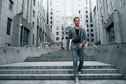 Laptop in hands. Young elegant man in good clothes is outdoors in the city at daytime.