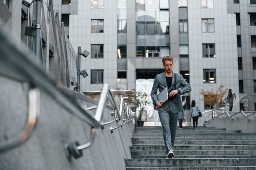 Laptop in hands. Young elegant man in good clothes is outdoors in the city at daytime.