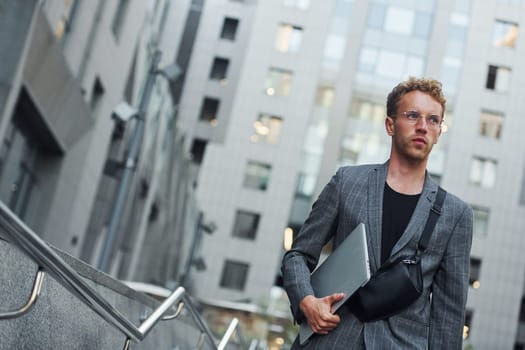 Laptop in hands. Young elegant man in good clothes is outdoors in the city at daytime.