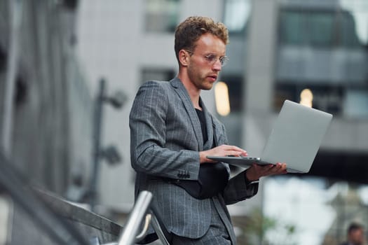 Laptop in hands. Young elegant man in good clothes is outdoors in the city at daytime.