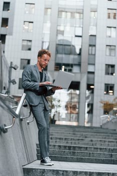 Laptop in hands. Young elegant man in good clothes is outdoors in the city at daytime.