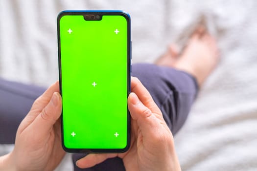 Woman hands hold a smartphone with green screen and sits on the bed background. Chroma key phone.