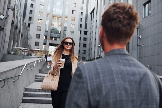 Girl holds drink. Woman and man in the town at daytime. Well dressed people.