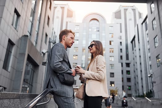 Girl holds drink. Woman and man in the town at daytime. Well dressed people.