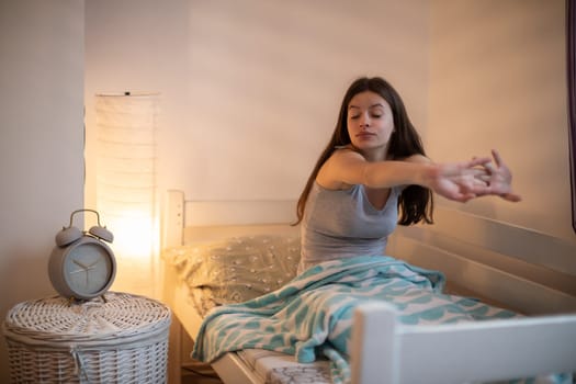 young woman stretches after sleeping. A large alarm clock stands on the bedside table.