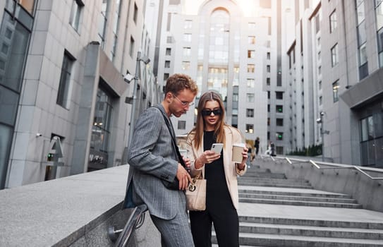 Girl holds drink. Woman and man in the town at daytime. Well dressed people.