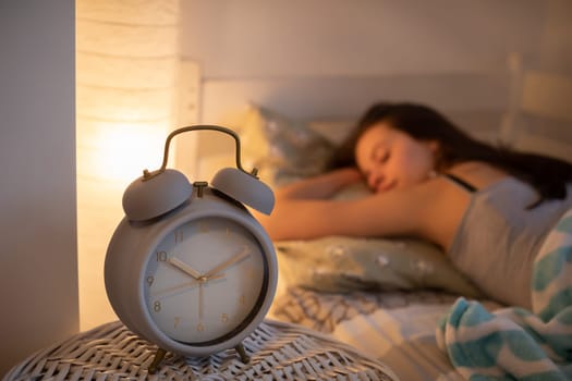 A young girl sleeps snuggled against her pillow. A large alarm clock stands on the bedside table.
