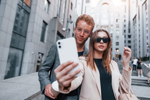 Makes selfie. Woman and man in the town at daytime. Well dressed people.