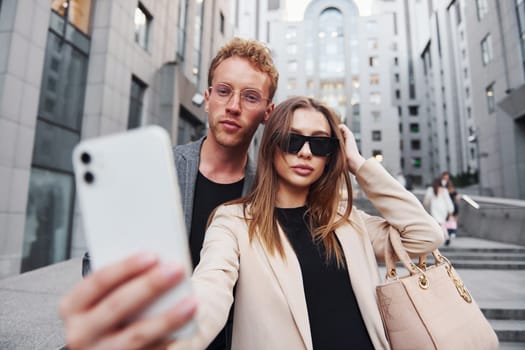 Makes selfie. Woman and man in the town at daytime. Well dressed people.