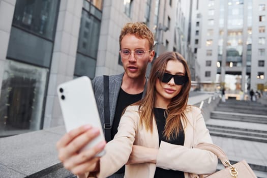 Makes selfie. Woman and man in the town at daytime. Well dressed people.