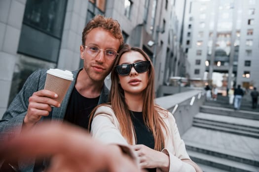 Makes selfie. Woman and man in the town at daytime. Well dressed people.