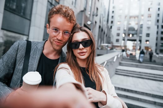 Makes selfie. Woman and man in the town at daytime. Well dressed people.