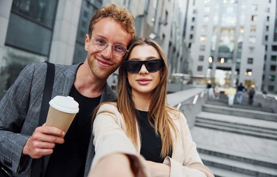 Makes selfie. Woman and man in the town at daytime. Well dressed people.