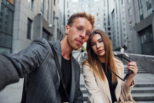 Makes selfie. Woman and man in the town at daytime. Well dressed people.