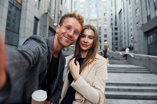 Makes selfie. Woman and man in the town at daytime. Well dressed people.