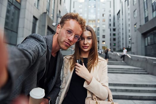 Makes selfie. Woman and man in the town at daytime. Well dressed people.