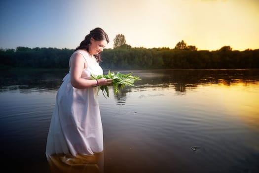 Slavic plump plump chubby girl in long white dress on the feast of Ivan Kupala with flowers and water in a river or lake on summer evening