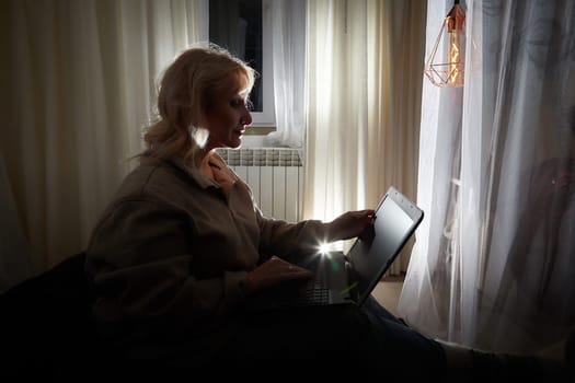 Silhouette and shadow of Adult mature woman of 40-60 years with laptop, notebook, computer in warm sweater in dark calm cozy evening atmosphere room. Interior with curtains and small linght