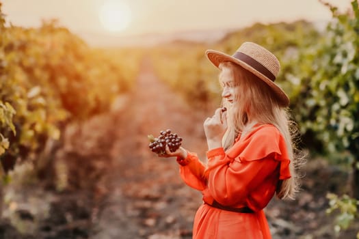 Portrait of a happy woman in summer vineyards at sunset. a woman in a hat and a red dress holds black grapes in her hands.