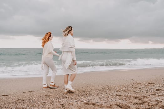 Women sea walk friendship spring. Two girlfriends, redhead and blonde, middle-aged walk along the sandy beach of the sea, dressed in white clothes. Against the backdrop of a cloudy sky and the winter sea. Weekend concept