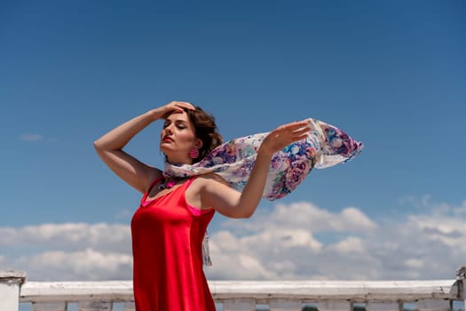 A woman in a red dress against a blue sky and white clouds with a developing scarf around her neck. She turned her face to the wind and posed for the camera