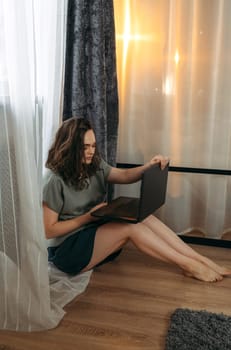 A beautiful woman is sitting on the floor near the window with a laptop, working or studying. Vertical frame.