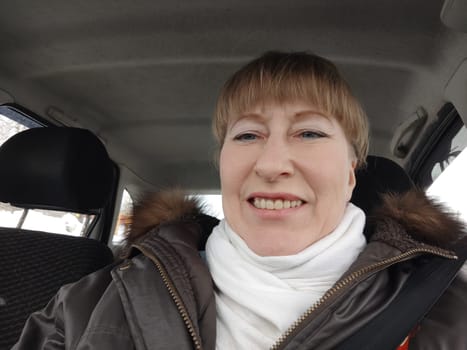 Portrait of adult cute funny blonde woman in warm clothes taking a selfie inside the car and driving it. The concept of pleasant trip in spring, autumn, winter