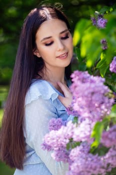 Happy brunette girl with long hair stands with lilac flowers, in the garden, in sunny day. Looking down. copy space