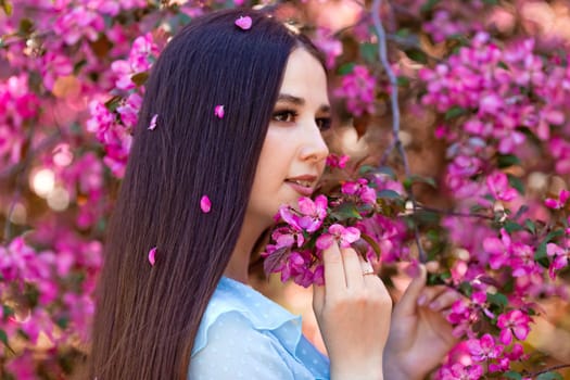 A brunette girl with pink petals in long hair is standing near a pink blooming apple tree, looking away. Close up.