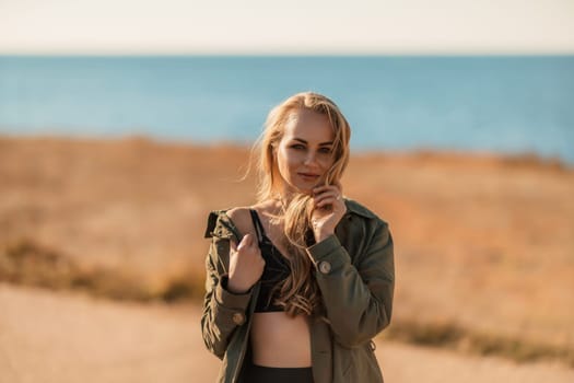 Portrait blonde sea cape. A calm young blonde in an unbuttoned khaki raincoat stands on the seashore, under the raincoat there is a black skirt and top.