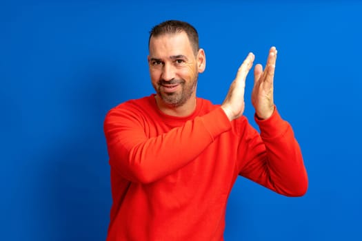 Bearded Hispanic man in his 40s wearing a red sweatshirt clapping excited about the great spectacle he just witnessed, isolated over blue background