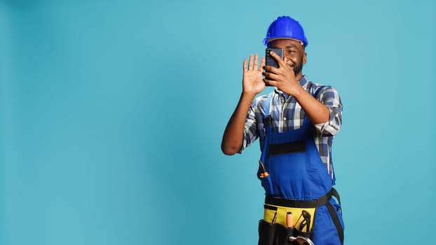 Cheerful builder talking on video call meeting in studio, using online videoconference chat to talk about new building contract. Young handyman in overalls using remote teleconference.