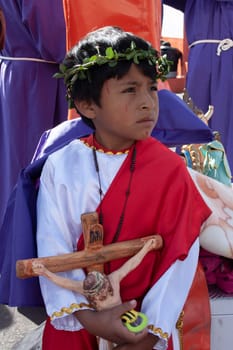 child in a profession of holy week dressed with a large cross of jesus christ in his arms with a sad look. from latam