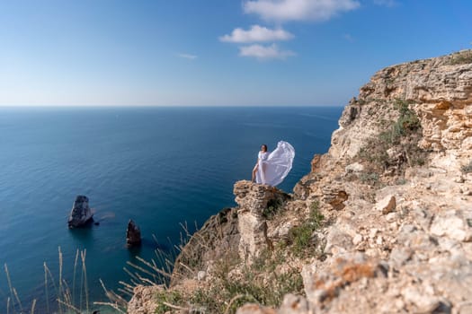 A beautiful young woman in a white light dress with long legs stands on the edge of a cliff above the sea waving a white long dress, against the background of the blue sky and the sea