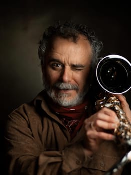 Confident and serious guy dressed in brown clothing poses in dark background holding saxophone as a weapon on his shoulders. High quality photo