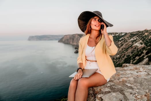 Successful business woman in yellow hat working on laptop by the sea. Pretty lady typing on computer at summer day outdoors. Freelance, travel and holidays concept.