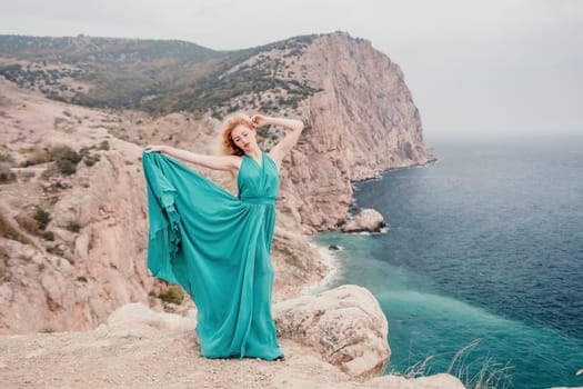Side view a Young beautiful sensual woman in a mint long dress posing on a volcanic rock high above the sea during sunset. Girl on the nature on overcast sky background. Fashion photo