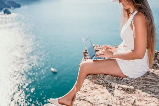 Successful business woman in yellow hat working on laptop by the sea. Pretty lady typing on computer at summer day outdoors. Freelance, travel and holidays concept.