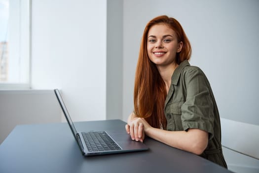 portrait of a cute, smiling red-haired woman sitting at a laptop with her hands folded on the table and looking at the camera. High quality photo