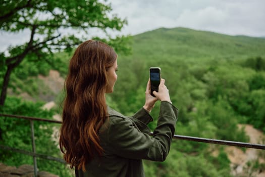 a woman in a raincoat takes pictures on the phone of a beautiful view of the forest. Travel, technology, mobile photography. High quality photo