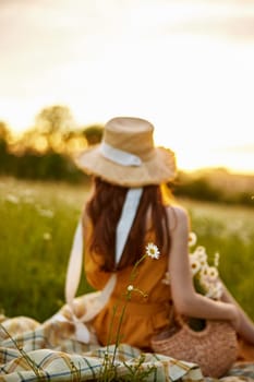 a woman in a wicker hat sits with her back to the camera in a chamomile field on a checkered plaid with a basket of flowers in her hands and looks at the sunset. High quality photo