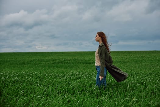 a beautiful woman stands in a green field in a raincoat and looks into the distance. Strong wind, flying hair, harmony with nature. High quality photo