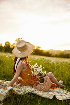 a woman in a wicker hat sits with her back to the camera in a chamomile field on a checkered plaid with a basket of flowers in her hands and looks at the sunset. High quality photo