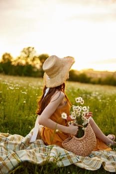 a woman in a wicker hat sits with her back to the camera in a chamomile field on a checkered plaid with a basket of flowers in her hands and looks at the sunset. High quality photo