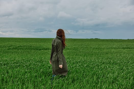 a red-haired woman in a long raincoat stands in a green field in cloudy weather with her back to the camera enjoying nature. High quality photo