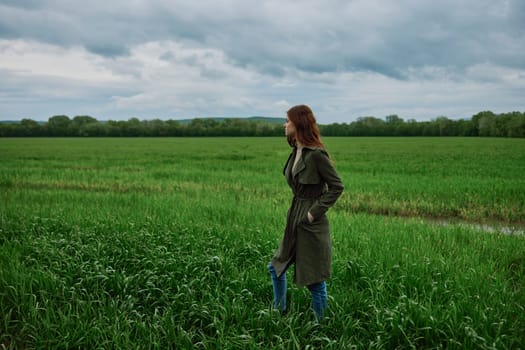 a woman with beautiful, long, red hair stands with her back to the camera in a green field in rainy, spring weather in a long raincoat. High quality photo