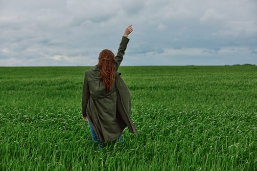 a red-haired woman stands with her back to the camera in a long coat with her hand raised up in a green field in rainy weather. High quality photo