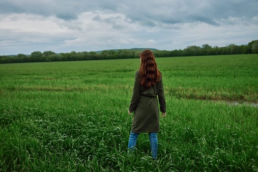 a woman with beautiful, long, red hair stands with her back to the camera in a green field in rainy, spring weather in a long raincoat. High quality photo