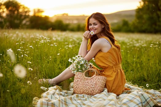 happy redhead woman sitting in a field of daisies on a plaid during sunset and smiling at the camera holding a flower in her hand. High quality photo